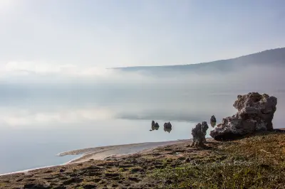 Early morning mist over the calm waters of Empakaai Crater Lake in Tanzania, with reflections, rocky shoreline and the forested volcanic crater illuminated by the rising sun.