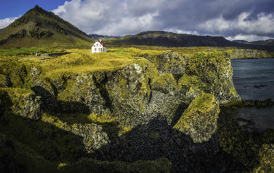 White farmhouse on grassy cliffs along the Snæfellsnes Peninsula in western Iceland, with rocky shoreline, ocean views, and mountains in the background.