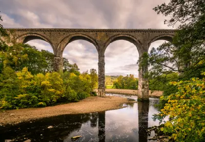 River-level view of Lambley Viaduct's tall stone arches reflected in the River South Tyne, surrounded by golden autumn trees and a rocky riverbank.