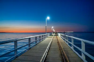 Railway tracks and illuminated walkway extending along Busselton Jetty over Geographe Bay at twilight.