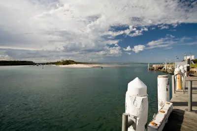 View across Currambene Creek from the Huskisson waterfront, with boats, a sandy shoreline and marina structures beneath a dramatic cloud-filled sky.