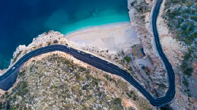 Top-down aerial view of Kaputaş Beach, showing a narrow sandy cove with turquoise Mediterranean water framed by rocky cliffs and a dramatically winding coastal road.