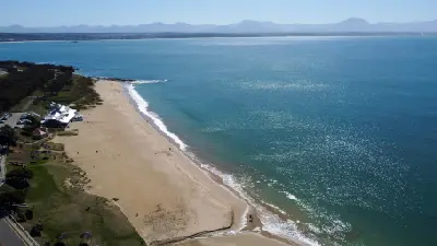 Aerial view of Santos Beach in Mossel Bay with calm turquoise water, a wide sandy shoreline and the Outeniqua Mountains in the distance.