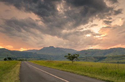 Road through grassland leading towards the Champagne Castle massif under dramatic sunset clouds in the Central Drakensberg, KwaZulu-Natal.