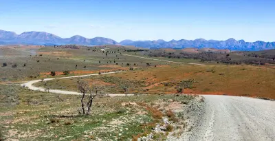 Winding gravel road through open plains and scattered vegetation in the Flinders Ranges, South Australia, with rugged mountain ranges stretching across the horizon.