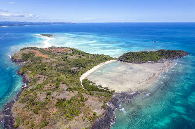 Drone view of Nosy Iranja, Madagascar, with volcanic island coastline, turquoise reef waters, white sandbanks, and the connecting tropical sandbar in the Indian Ocean.