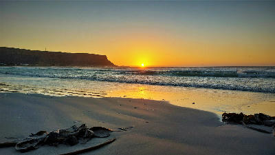 Sun setting beside Baboon Point at Elands Bay, with golden reflections on wet sand, gentle surf and the sandstone headland silhouetted against the evening sky.
