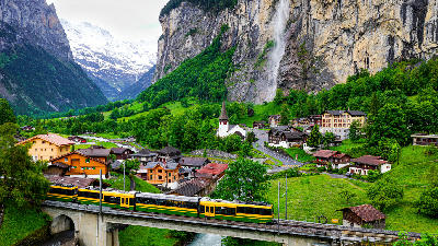 Train crossing a bridge in Lauterbrunnen village with Staubbach Falls on a cliff and alpine valley landscape in Switzerland