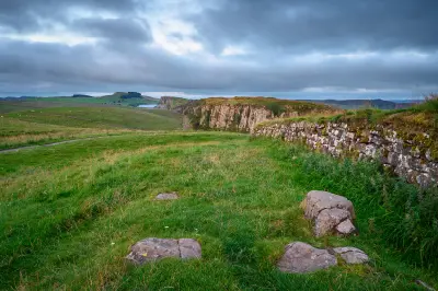 Hadrian's Wall beside grassy moorland at Steel Rigg, with the rocky cliffs of Peel Crags stretching into the distance beneath heavy clouds in Northumberland, England.