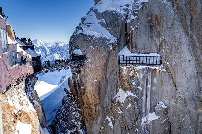 Visitors crossing a bridge at Aiguille du Midi high above the Mont Blanc Massif with surrounding alpine peaks.
