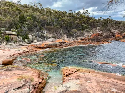 Clear water surrounded by pink granite rocks, orange lichen and coastal forest at Sleepy Bay, Freycinet National Park, Tasmania.