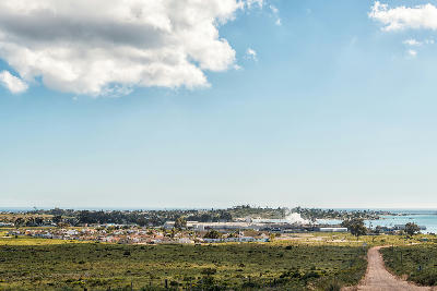 Elevated view of St Helena Bay with coastal town, harbour facilities, open grassland and the Atlantic Ocean stretching across the horizon under a partly cloudy sky.