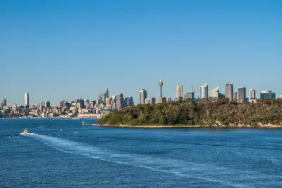 Forested Bradleys Head peninsula extending into Sydney Harbour, with the CBD skyline beyond and boats crossing the blue harbour waters.