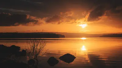 Golden-orange sunset over Kielder Water in Northumberland, with the sun reflecting on calm reservoir water, dark clouds overhead and silhouetted rocks in the foreground.