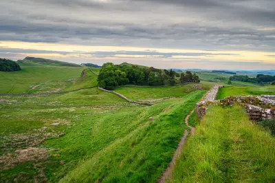 Hadrian's Wall crossing green hills near Housesteads Roman Fort in Northumberland, with woodland, distant ridges and layered evening clouds.