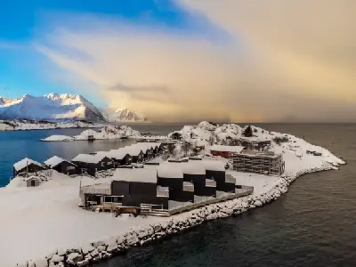 Elevated view of black rorbu cabins and harbour at Hamn i Senja surrounded by snow-covered rocky shoreline and open sea