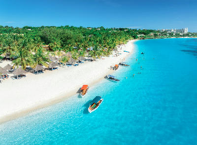 Drone view of Kendwa Beach showing crystal-clear turquoise water, anchored boats, white sand and palm-lined shoreline on Zanzibar's north coast, Tanzania.