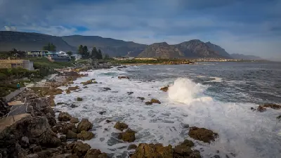 Panoramic view of the Hermanus Cliff Path with rugged coastal rocks, crashing ocean waves, Walker Bay, and mountain scenery along the Whale Coast in the Western Cape, South Africa.