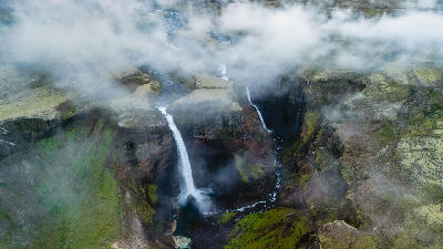 Aerial view of Háifoss waterfall in the Iceland Highlands, cascading into a narrow canyon with green moss, rocky cliffs, and low cloud cover.