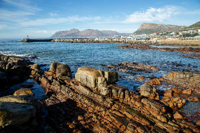 Rocky coastline and harbour at Kalk Bay with layered sandstone rocks, breakwater, and coastal town along False Bay in Cape Town, South Africa.