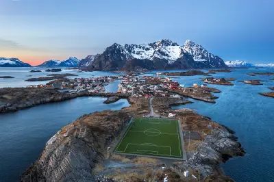 Drone view of Henningsvær on Austvågøya in the Lofoten Islands, showing the coastal football field on a rocky islet with harbour settlement and snow-covered peaks behind in Nordland, Norway.