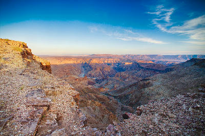Cliff-edge sunrise view across Fish River Canyon in southern Namibia showing layered canyon walls, deep river valleys, and expansive desert escarpments beneath a blue sky.