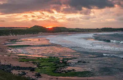 Dramatic orange sunset over Embleton Bay in Northumberland, with breaking waves, wet sandy shoreline, seaweed-covered rocks and coastal dunes.