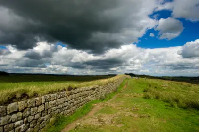 Hadrian's Wall stretching into the distance at Steel Rigg in Northumberland, England, with walkers on a grassy path beneath dramatic dark storm clouds.