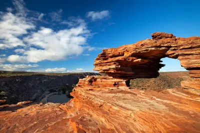Nature’s Window rises above a broad red sandstone platform overlooking the Murchison River gorge beneath a blue, cloud-filled sky.