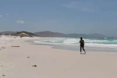 Lone person walking along the long white-sand shoreline of Friendly Beaches, with turquoise waves and distant coastal hills in Freycinet National Park, Tasmania.