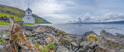 White church of Viðareiði on a stone wall above the rocky fjord coast with distant islands and cloud-covered mountains in the Faroe Islands.