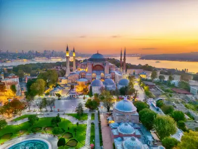 Aerial view of Hagia Sophia and Sultanahmet Square in Istanbul at sunset, with illuminated minarets, gardens, the Bosphorus and city skyline.