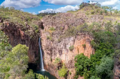 Elevated view of Tolmer Falls dropping between tall orange sandstone cliffs into a dark plunge pool surrounded by tropical woodland in Litchfield National Park, Northern Territory, Australia.