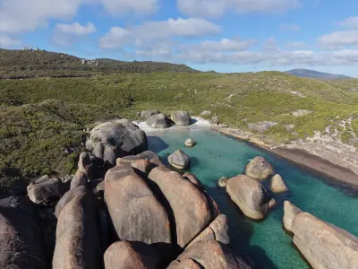 High-angle aerial view of a sheltered cove featuring massive rounded granite boulders resembling elephants, clear turquoise water with swimmers, a white sand beach, and lush green coastal hills under a blue sky.