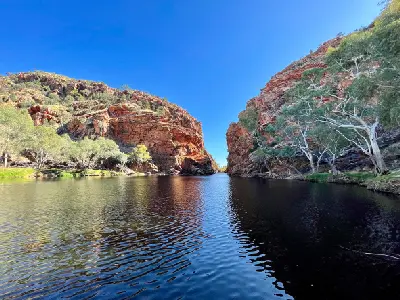 Wide waterhole cut through a desert gorge with sheer red cliffs and sparse bush under a deep blue sky in the Northern Territory.