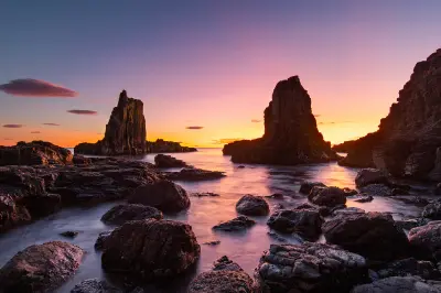 Cathedral Rocks rise from the ocean beneath a vivid pink and orange dawn sky, with long-exposure water flowing around the rocky shoreline.
