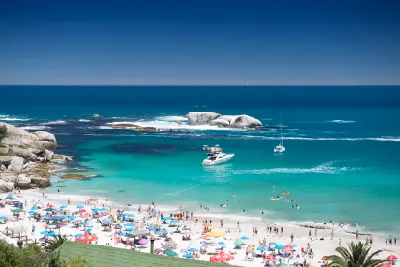 Busy summer day at Clifton Beach in Cape Town showing beach umbrellas, swimmers in clear turquoise water and yachts anchored offshore near large granite boulders.