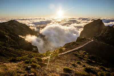 Sunset over the mountain ridges near Pico do Arieiro, with a stone path leading through high terrain above a sea of clouds.