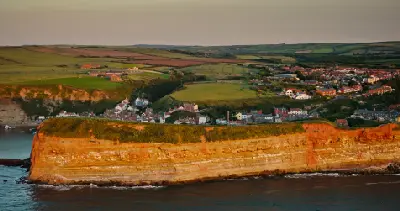 Aerial view of sunlit sandstone cliffs, Staithes fishing village and rolling farmland along the North Yorkshire coast, England.