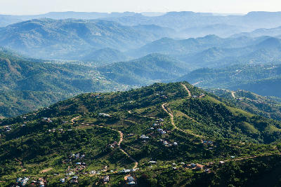 Elevated view of the Valley of a Thousand Hills in KwaZulu-Natal, showing layered green hills, winding roads, rural homesteads and distant mountain ridges under hazy light.