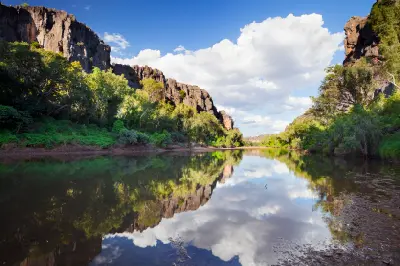 Still water reflects limestone cliffs, lush riverside vegetation and white clouds inside Bandilngan (Windjana Gorge).