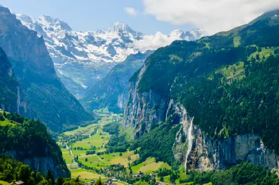 Elevated view over Lauterbrunnen Valley from Wengen, showing green valley floor, steep cliff walls with waterfalls, and snow-capped peaks in the background, Switzerland.