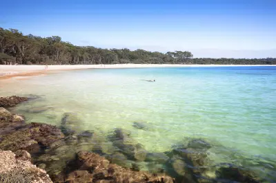 Clear emerald water laps against rocks and a sweeping white-sand shoreline backed by forest at Green Patch Beach.