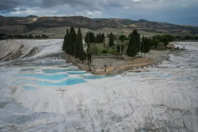 Panoramic view of Pamukkale’s white travertine terraces and pale blue thermal pools, surrounded by cypress trees and mountains beneath a cloudy sky.