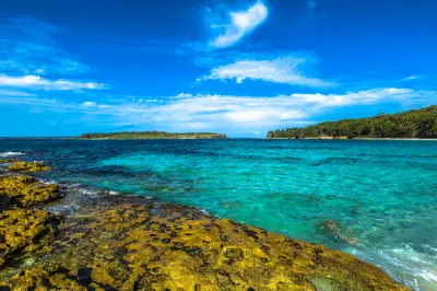 Clear turquoise water meets a rocky shoreline at Hyams Beach, with forested headlands extending across Jervis Bay.