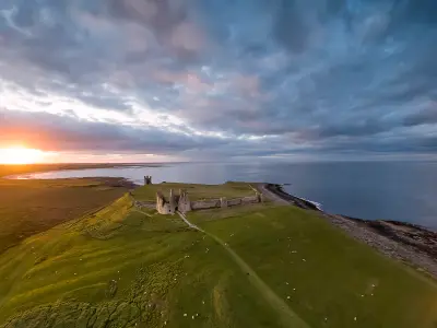 Aerial view of Dunstanburgh Castle on a grassy coastal headland at sunset, with Embleton Bay, the North Sea and dramatic clouds over Northumberland.