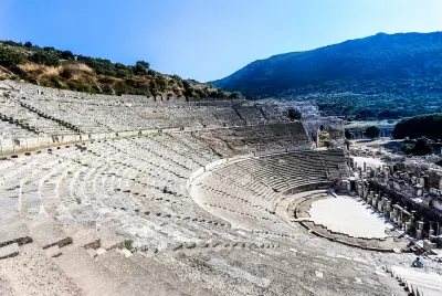 Curving tiers of ancient stone seats descend toward the stage of the Great Theatre of Ephesus, with a wooded hillside rising behind the ruins.