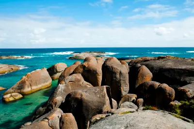 Clusters of large, smooth granite boulders line a clear turquoise coastal inlet under a bright blue sky with light clouds.