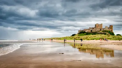 Bamburgh Castle overlooking a sandy Northumberland beach, with visitors walking along the shoreline, reflections on wet sand and dark storm clouds overhead.