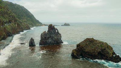 Rocky coastline with volcanic sea stacks and a natural rock arch at Ribeira da Janela, Madeira, Portugal, backed by steep green cliffs and the Atlantic Ocean.
